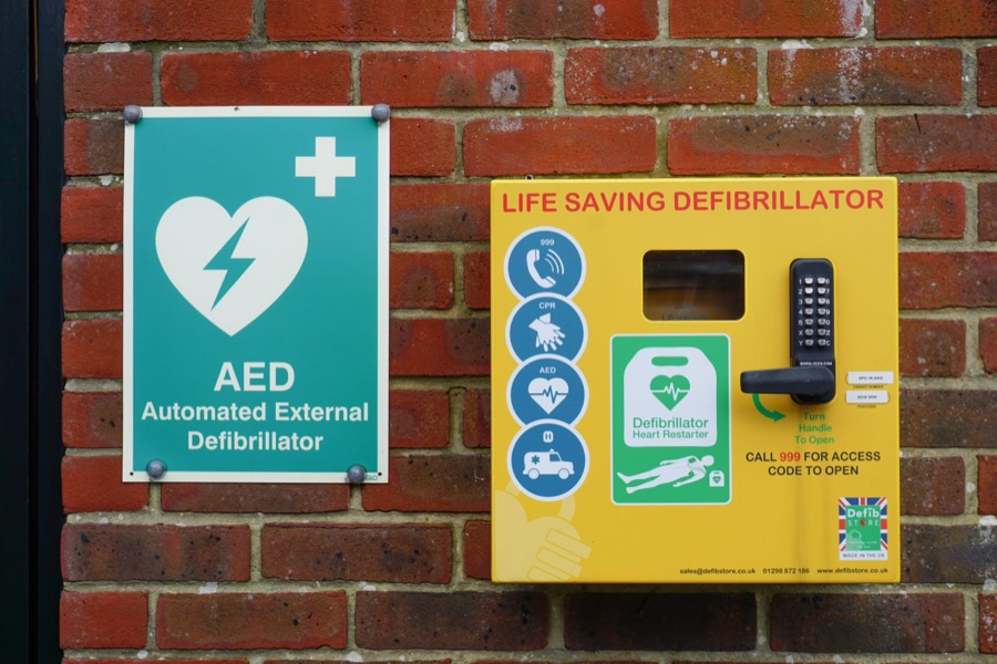A public defibrillator: bright yellow wall cabinet with a black keypad lock, next to a green AED sign